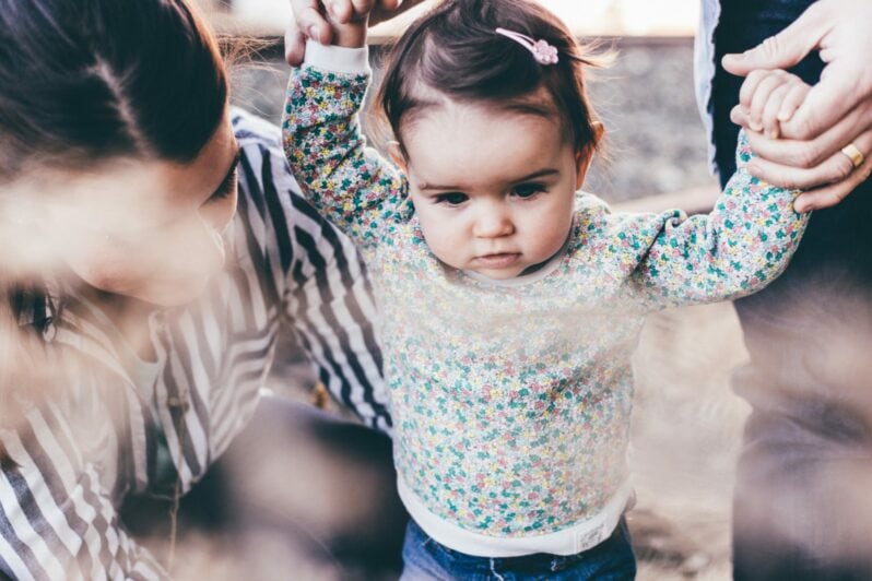 Caregivers talk to a young child who is learning to walk.