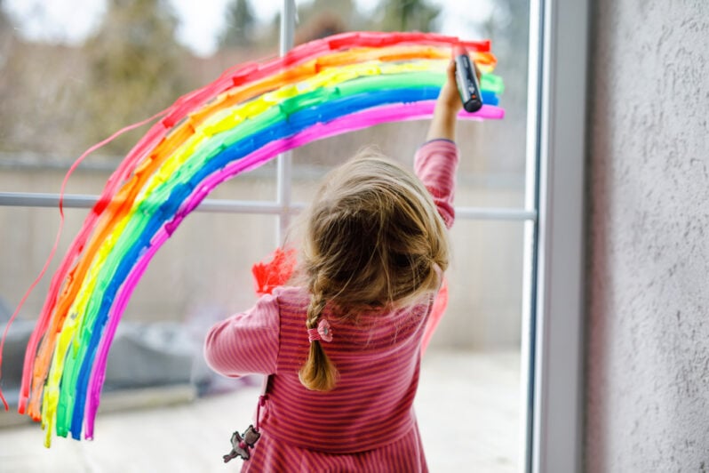 A young girl paints a rainbow on a window.