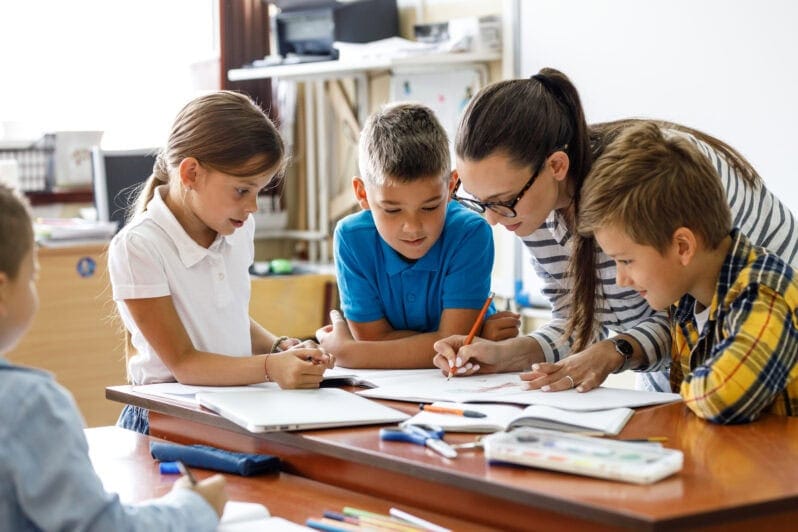 Children huddle round a teacher who is writing on paper.