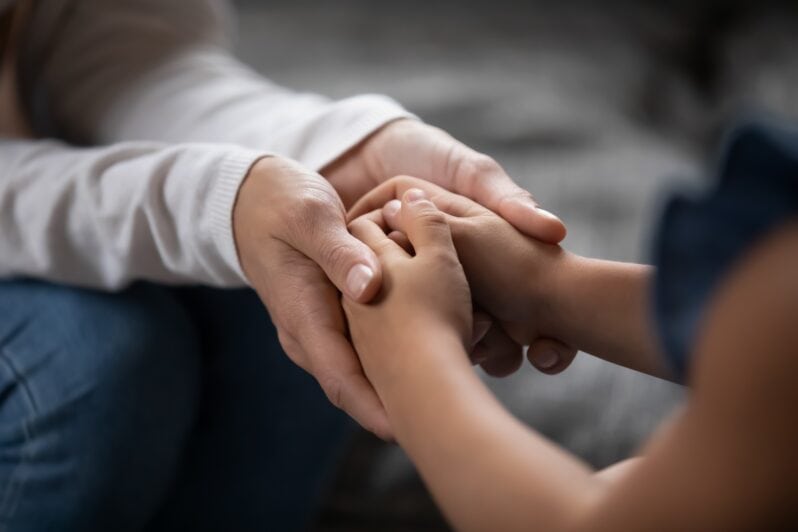 An adult sits opposite a child, holding their hands for comfort.
