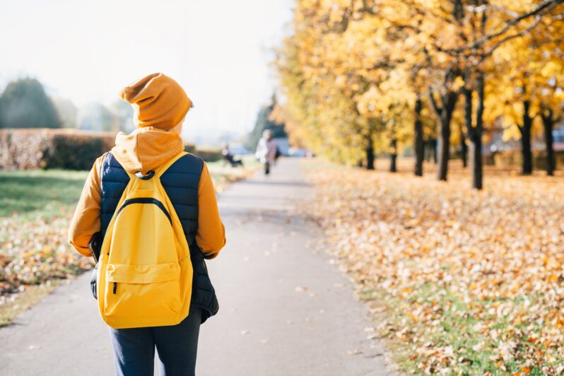A young person wearing a backpack walks along a tree-lined path.