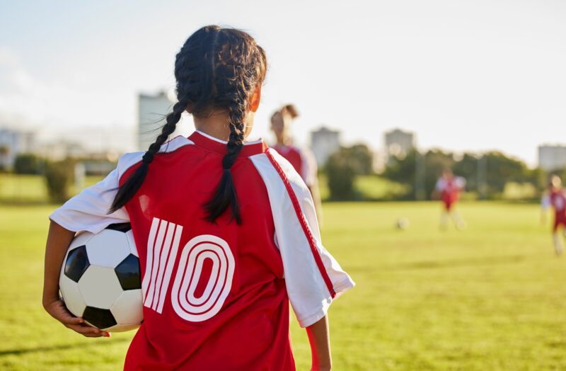 A girl looking away from the camera holding a football on a football pitch