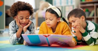 Three children are lying on the floor, reading a book together.
